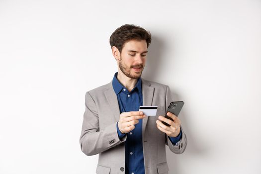 Online shopping. Smiling businessman paying with credit card on smartphone, sending money, standing in suit on white background.