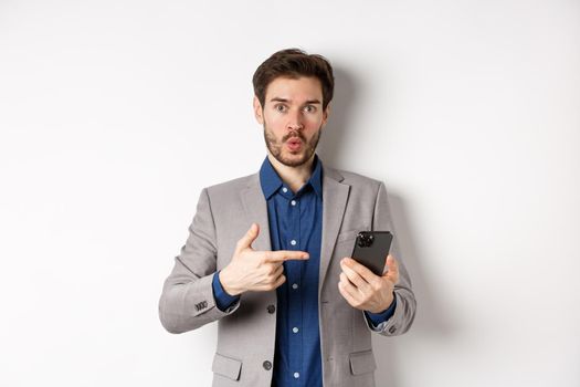 Excited guy in suit say wow, found something online on smartphone, pointing at mobile phone impressed, white background.