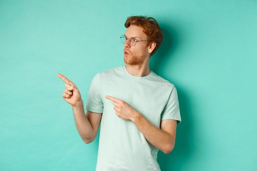 Impressed redhead man in glasses and t-shirt, pointing fingers and looking left at promo offer, staring happy, standing over turquoise background.