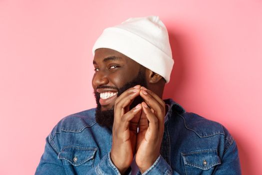 Close-up of devious african-american male model having an idea, scheming something, steeple fingers and smiling sly, standing over pink background.