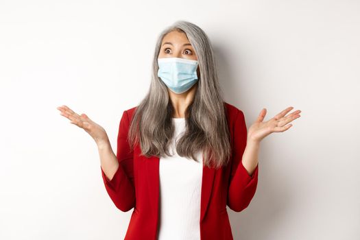 Coronavirus and business concept. Surprised asian businesswoman in face mask, spread hands sideways and staring shocked at upper left corner, white background.