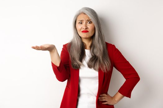 What is this. Confused and skeptical asian female entrepreneur pointing hand at something strange, staring at camera puzzled, standing over white background.
