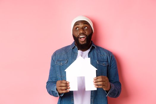 Real estate concept. Handsome african-american man dreaming about home, holding house model and looking at upper left corner, imaging apartment, pink background.