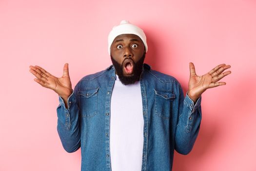 Shocked and impressed Black man staring at camera with complete disbelief, saying wow, standing in beanie and hipster shirt over pink background.