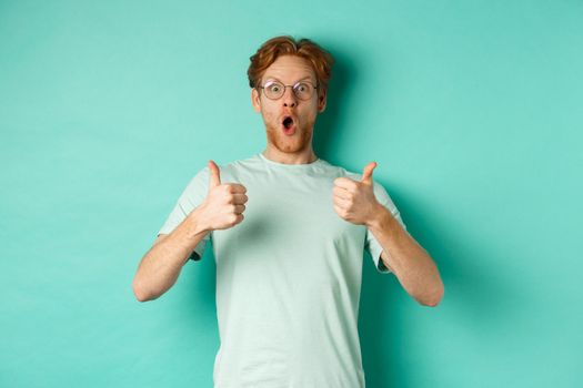 Amazed young man with red hair and beard, wearing glasses with t-shirt, showing thumbs-up and gasping in awe, checking out awesome promo offer, turquoise background.