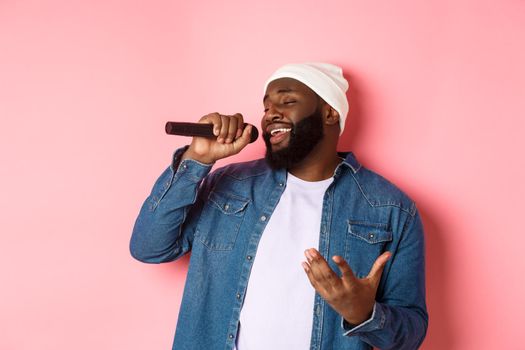 Handsome Black man in beanie and denim shirt singing karaoke, holding microphone, standing over pink background.