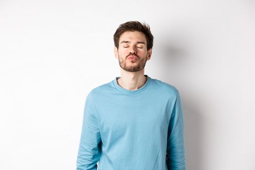 Attractive boyfriend waiting for kiss with puckered lips and closed eyes, standing in sweatshirt over white background.