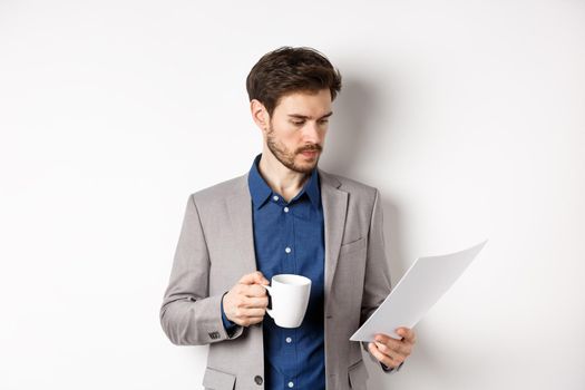 Boss reading documents and drinking morning coffee from mug, looking at paper serious, standing in suit on white background.