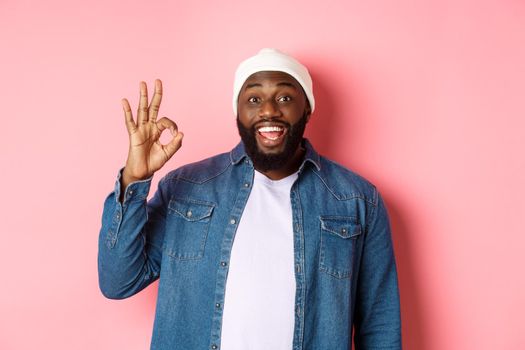 Happy smiling Black guy showing okay sign, approve and praise good offer, standing over pink background.