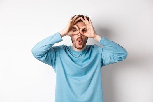 Check it out. Impressed young man seeing awesome promo, looking through hand binoculars and saying wow, standing over white background. Copy space