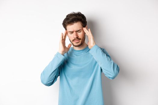 Young man having headache or hangover, touching head temples and frowning, feeling sick from migraine, standing over white background .