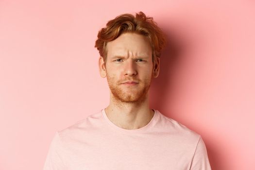 Close up of suspicious redhead guy frowning, staring at camera doubtful, express disapproval, standing over pink background.