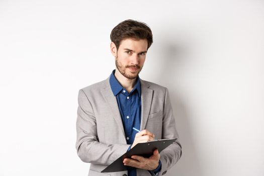 Successful businessman in grey suit taking notes, working in office clothing, holding clipboard and looking at camera, white background.