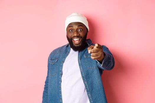 Happy african-american hipster man pointing finger at camera, need you, smiling excited, standing over pink background.