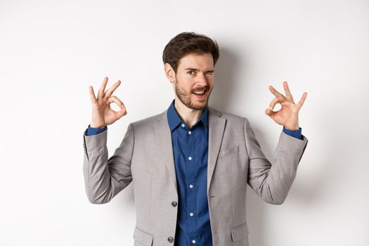 Cheeky handsome man in suit winking at you, showing okay signs, all under control gesture, approve good choice, assuring everything OK, standing on white background.