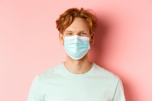 Covid-19 and pandemic concept. Close up of redhead man in medical mask looking at camera, standing over pink background.