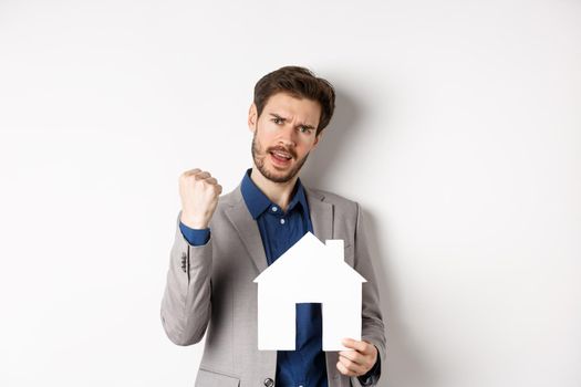 Real estate. Excited man buying house and showing paper home cutout, say yes and fist pump with joy, standing in suit on white background.