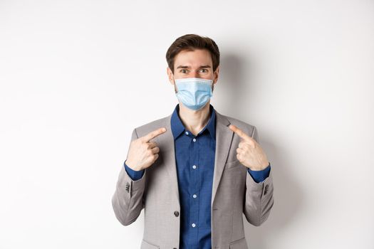 Covid-19, pandemic and business concept. Excited businessman in suit pointing at his medical mask and smiling with eyes, white background.