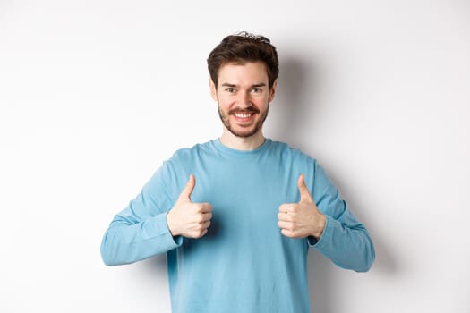 Smiling handsome man showing thumbs up, like and approve good choice, praising or recommending something, standing on white background.