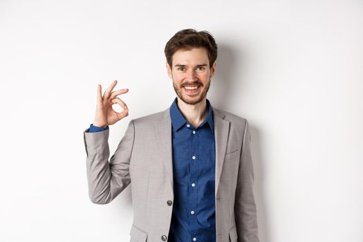 Smiling successful businessman in suit showing okay sign, looking satisfied, standing against white background.