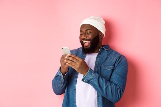 Technology and online shopping concept. Happy Black bearded man reading message and smiling, using smartphone against pink background.