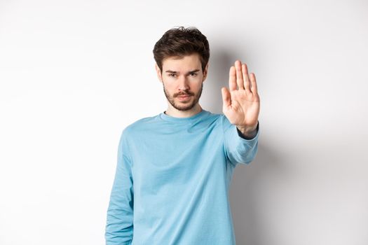 Serious young man looking confident with hand stretched out, saying stop, prohibit something bad or give warning, standing over white background.