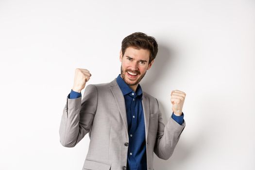 Lucky businessman winning money prize, say yes and smiling excited, make fist pump sign to celebrate victory, triumphing in suit on white background.