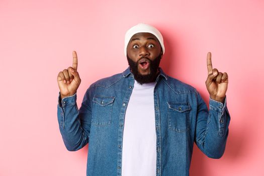 Excited and impressed Black man showing amazing promo, pointing fingers up at logo, standing against pink background.