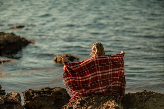 Attractive blonde Caucasian woman enjoying time on the beach at sunset, sitting in a blanket and looking to the side, with the sunset sky and sea in the background. Beach vacation