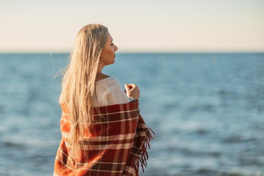 Attractive blonde Caucasian woman enjoying time on the beach at sunset, walking in a blanket and looking to the side, with the sunset sky and sea in the background. Beach vacation.