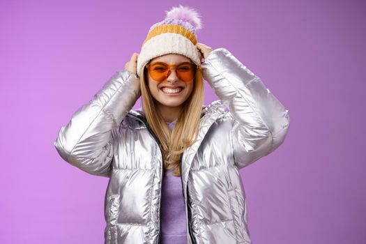 Energized daring sassy young attractive woman having fun friends winter trip learn snowboarding smiling cheeky enjoying vacation put-on hat wearing silver warm jacket sunglasses, purple background.
