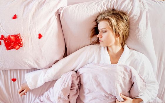 Happy valentines day. young blond woman sleeping in bed with holiday gift and red confetti