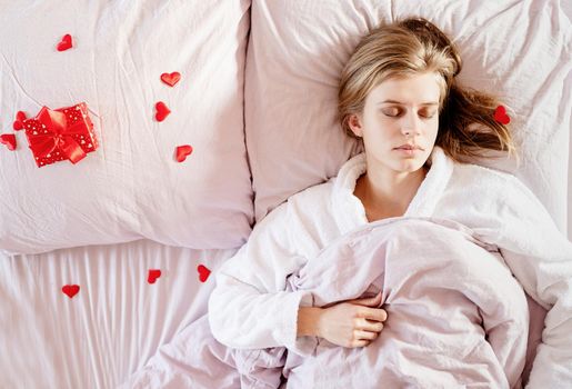 Happy valentines day. young blond woman sleeping in bed with holiday gift and red confetti