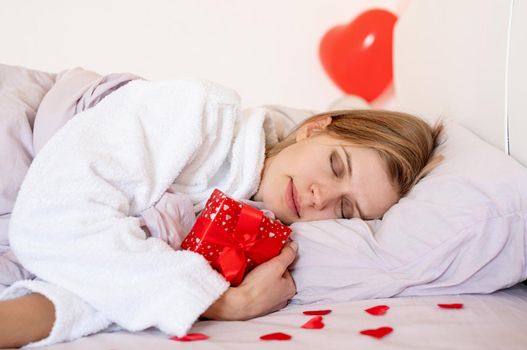 Happy valentines day. young blond woman sleeping in bed with holiday gift and red balloons