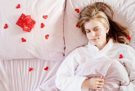 Happy valentines day. young blond woman sleeping in bed with holiday gift and red confetti