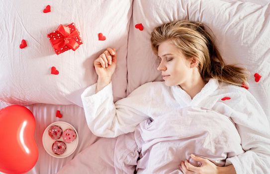 Happy valentines day. young blond woman sleeping in bed with holiday gift and red confetti