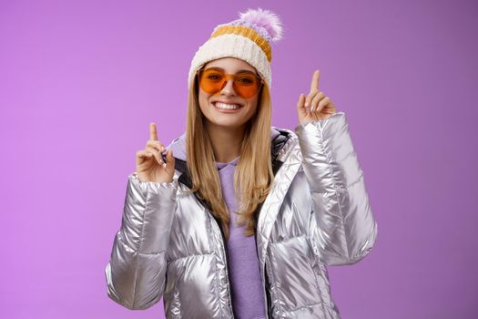 Joyful energized entertained cute blond woman having fun enjoy vacation snowy mountain trip wearing sunglasses silver jacket winter hat dancing pointing up amused standing purple background.