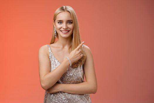 Confident attractive feminine glamour blond young woman in silver glittering dress smiling broadly pointing up discuss recent news promo sales standing friendly grinning camera red background.