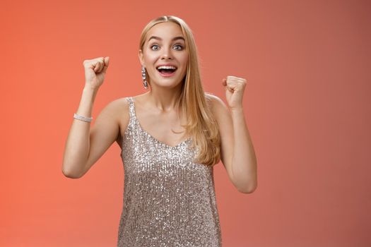 Surprised happy celebrating blond young woman in silver trendy dress raising hands up yes victory gesture smiling broadly excited winning first place reach goal grinning thrilled triumphing.