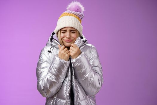 Unhappy sobbing whining cute blond girl pull jacket tight body close eyes crying freezing cold standing snowy winter resort shaking low temparature, purple background suffering discomfort.
