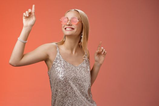 Delighted carefree attractive stylish millennial blond woman celebrating partying having fun wear sunglasses trendy silver dress dancing closed eyes broad smile waving hands up, red background.