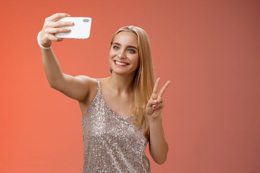 Attractive feminine tender young blond girl 25s in silver stylish dress taking selfie extend arm up show peace gesture smartphone display record video greeting internet fans, standing red background.