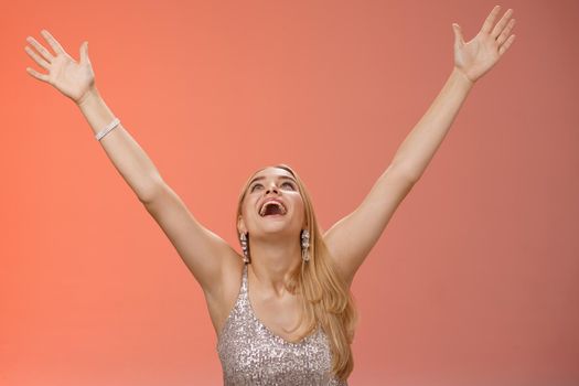 Happy emotive smiling overwhelmed young blond woman in silver dress raise hands sky thank god joyfully signed contract got job rejoicing red background celebrating victory good news, triumphing.