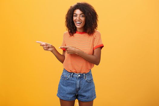 Ask him. Portrait of friendly and joyful good-looking stylish female shop assistant with curly hair and dark skin pointing left with both hands, smiling assured and entertained over orange background.