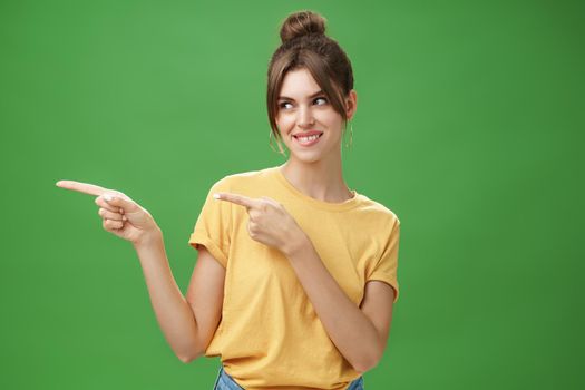 Waist-up shot of charismatic happy and carefree charming woman in yellow t-shirt pointing and looking left enthusiastic and pleased smiling cheerfully posing against green background delighted. Copy space