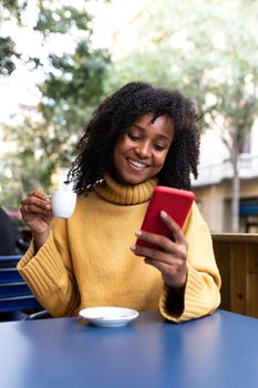 Happy young African American woman using mobile phone and drinking coffee in an outdoors coffee shop terrace. Vertical image. Lifestyle and technology concept.