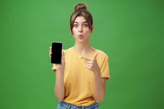 Portrait of intrigued woman cannot wait use new smartphone holding phone and pointing at cellphone screen folding lips with excitement and interest presenting new device model in store over green wall. Lifestyle.
