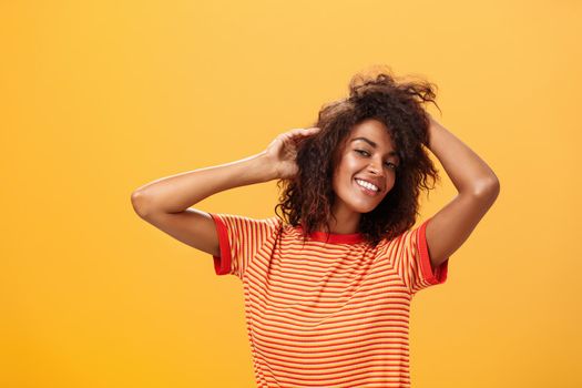 Waist-up shot of charming flirty feminine dark-skinned female in playful mood dancing playing with curly hair and smiling with delight and joy posing over orange background happy and carefree. Lifestyle.