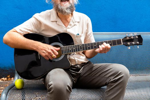 Unrecognisable mature man with beard playing the guitar and singing sitting on a bench on blue background. Musical instrument practice.