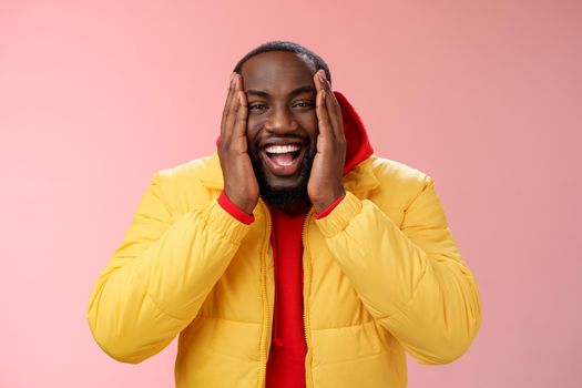 Happy charismatic playful african american bearded guy in yellow jacket red hoodie press palms cheeks smiling friendly laughing happily joking cannot believe receive awesome surprise, winning lottery.